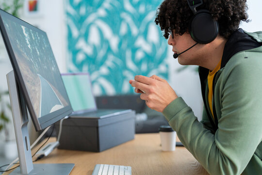 Side view of Arab young male in hoodie and headset holding gamepad while playing videogame on computer at desk in cozy room