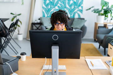 curly guy sitting in front of monitor of computer with laptop aside and playing video game with gamepad at home