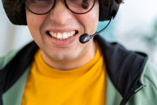 Crop young Arab male in eyeglasses and headset with microphone smiling while playing game and spending time at home