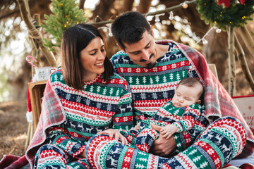 Parents in matching festive pajamas cuddling their infant outdoors, cozy holiday bonding and joyful new-parent portrait in winter forest