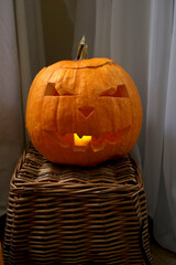 Рumpkin with a candle in the middle on a wicker stand made of vines on a background of fabrics. A scary face is carved on the pumpkin. Halloween theme, holiday decorations, decoration, celebration