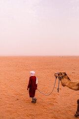 Moroccan man in traditional red robe and white headscarf leading a camel through the golden desert sands of the Sahara under bright sunlight, symbolizing nomadic life and Moroccan culture.