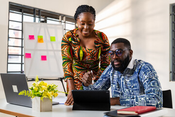 Two African corporate professionals collaborate in a modern office, sharing ideas and working...