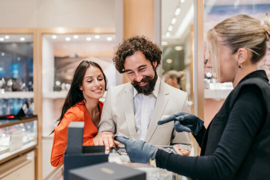 Excited man with his smiling partner are interacting with a sales assistant in a retail store, looking at high-end watches displayed on the counter