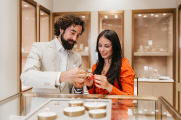 Young happy couple standing at a display counter, looking at different luxurious rings and trying on jewelry, celebrating an important life milestone in a high-end retail environment