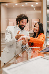 Obraz premium Young elegant couple tries on luxury wristwatch or bracelet in upscale jewelry store, smiling as they choose the perfect timepiece with attentive sales assistance and refined atmosphere