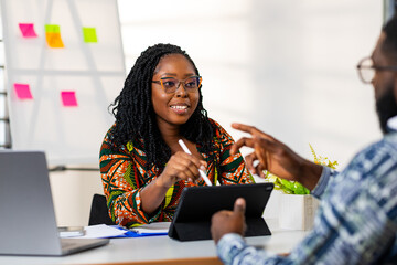 African businesswoman in vibrant ankara dress engages in a lively discussion with a colleague, using a tablet in a modern office setting