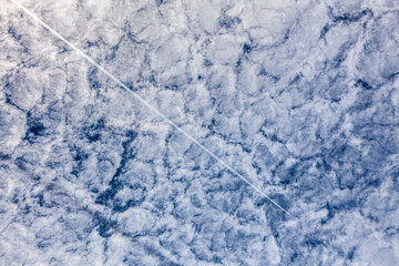 A straight aircraft contrail slices through a mottled sky of altocumulus clouds—highlighting contrast, motion, and atmospheric geometry.