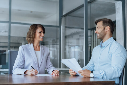Mature woman client talking with young man bank real estate manager worker in office. Two business entrepreneur people sitting at table signing contract document at meeting, discussing cv on interview