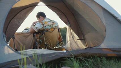 Romantic back view of an Asian couple sitting together inside a tent, silently watching the tranquil sunset over the lake during their camping date - Powered by Adobe