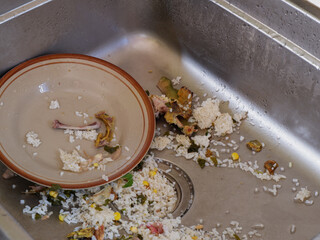 Dirty kitchen sink with leftover food scraps, rice, and bones on a plate. Messy dishwashing area showing unclean dishes and food waste after a meal, symbolizing household chores and cleaning.