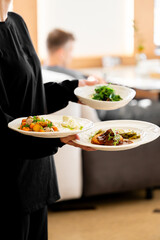 Waiter in black uniform serving three gourmet dishes in a modern restaurant setting. Plates feature salad, roasted vegetables, and meat with sauce. Indoor dining atmosphere.