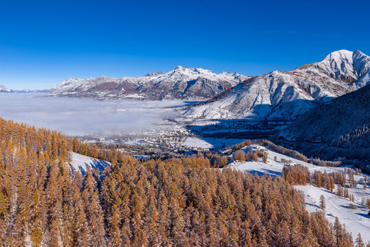 Aerial autumn view of the Champsaur Valley in the French Alps, with snow-dusted golden larch forest and mountains. The village of Ancelle emerges from morning fog. Hautes Alpes, France