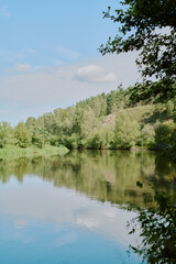 Fototapeta premium Calm river reflecting green trees and blue sky with scattered clouds, forested hillside rising in background, leafy branches framing scene, tranquil natural landscape, no people visible