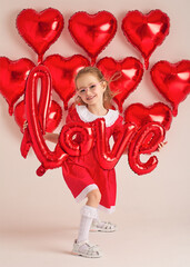 Smiling girl in red dress holds shiny balloon word &ldquo;love&rdquo;.
Heart balloons create festive Valentine background.
Playful studio portrait captures childhood joy and romance theme.