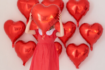 Little girl in red polka-dot dress hides face behind heart balloon.
Creative Valentine studio concept with festive red decor.
Playful portrait shows childhood joy and romantic mood.
