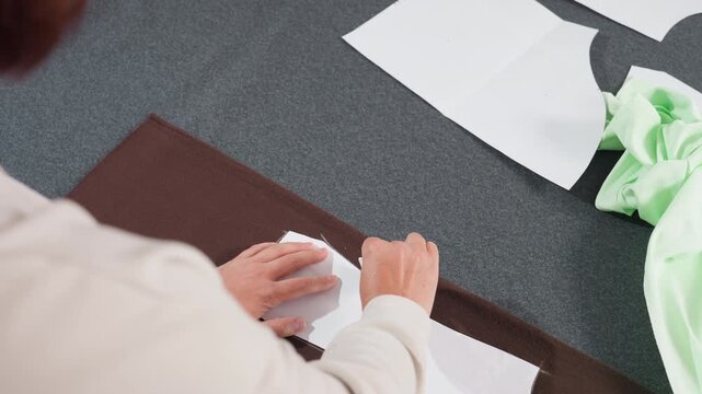 Garment designer sketching pattern with pencil on white paper sheet placed over dark brown fabric while working carefully in tailoring workshop surrounded by scattered sewing materials