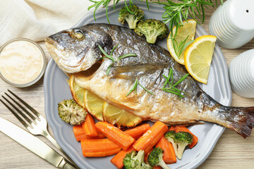 Cooked fish served with rosemary, lemon and vegetables on wooden table, flat lay