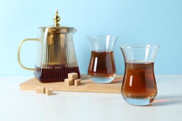 Tasty Turkish tea in glass cups, teapot and brown sugar on white marble table against light blue background