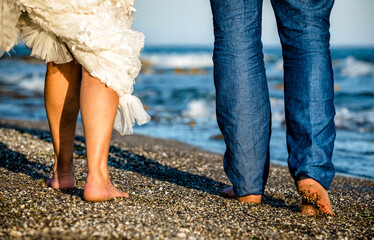 Barefoot couple walking on pebbled beach in sunlight wearing wedding dress and blue trousers symbolizing love, togetherness, freedom, honeymoon, and romantic coastal lifestyle.