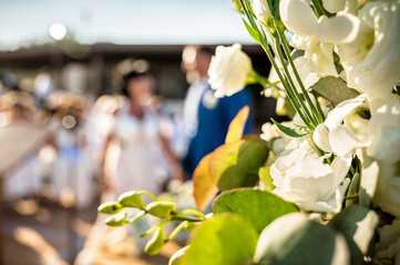 Close-up of floral wedding arch with white flowers and green leaves, blurred couple standing during ceremony in sunlight. Romantic moment captured with artistic composition.
