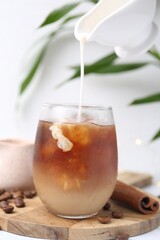 Pouring milk into tasty iced coffee in glass on white table, closeup