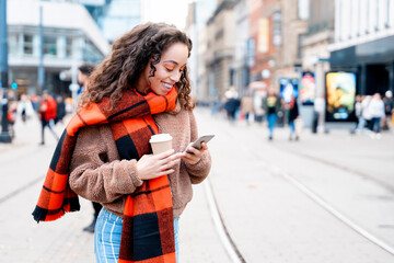 Woman smiles while using smartphone on busy city street during daytime in autumn with warm beverage in hand