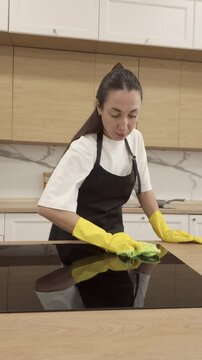 Young woman in black apron and yellow gloves wiping stove surface with green cloth in modern kitchen. Bright daylight, clean interior, concept of hygiene and home care