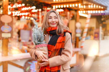 Woman holding miniature Christmas tree at festive market wearing warm scarf during holiday season