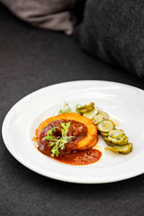 Gourmet dish with polenta cake, meat in red sauce, and herb-garnished cucumber slices on a white plate. Elegant restaurant-style plating on dark fabric background.