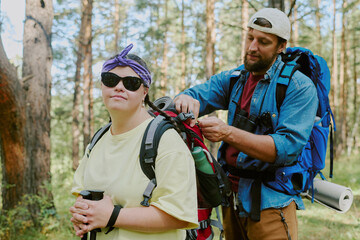 Caucasian young girl with Down syndrome wearing sunglasses standing in forest holding cup, while Caucasian young man adjusting backpack straps outdoors during hiking trip in summer woodland
