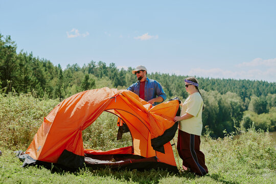 Caucasian young adult man and young girl with Down syndrome assembling orange tent outdoors on grassy hillside with forest in background, both focused on setting up camping equipment together