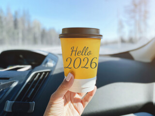 Hand holding yellow paper coffee cup with inscription Hello 2026 inside car, snowy road winter landscape visible outside window, concept of travel, new beginnings, adventure modern lifestyle © Evgeniia Primavera