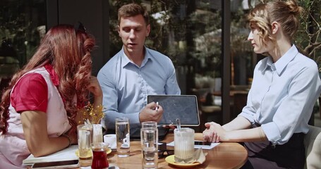 Three business partners engage in a productive outdoor meeting, using a tablet for presentation. They are collaborating, discussing strategies, and sharing ideas in a relaxed cafe setting. - Powered by Adobe