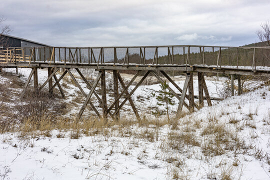Wooden Footbridge over Snowy Landscape with Cloudy Sky and Distant Hills. Winter scenery with bare trees, grasses, and distant hills beneath a gray, cloudy sky. - Powered by Adobe