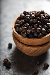 Whole black peppercorns in wooden bowl on light grey surface vertical shot, sharp focus with shallow depth of field.