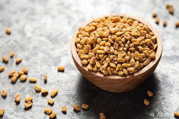 Fenugreek whole seeds in wooden bowl on grey surface with scattered grains around. Often used in...