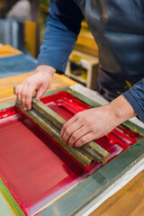 Detail of hands working with screen printing frame and red ink on fabric. Artisan printing process in workshop. Traditional technique. Creative and authentic mood.