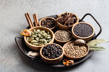 Ingredients for preparing traditional Indian garam masala whole aromatic spices in wooden bowls on rustic black plate on grey background, traditional Indian cooking concept.