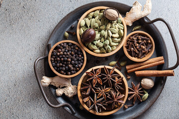 Composition of ingredients for traditional Indian masala chai on black tray, featuring whole aromatic spices such as cardamom, cinnamon, cloves, black pepper, ginger on rustic background.
