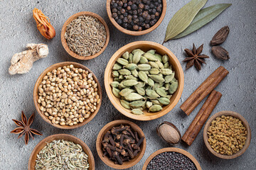 Ingredients for preparing garam masala whole aromatic spices in wooden bowls on grey background, close-up top view, traditional Indian cooking and rich natural textures.
