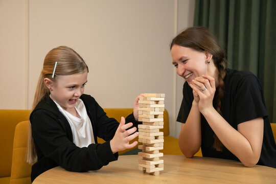 Little girl concentrates as she carefully pulls a wooden jenga block from the tower while her mother watches nearby with a calm, supportive smile during cozy game time at home