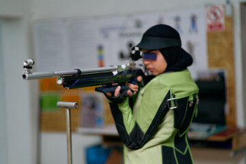 Female athlete wearing professional shooting uniform and hijab carefully aiming with an air rifle at an indoor range.