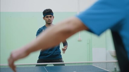 Young man in blue shirt prepares to return shot during intense indoor table tennis match, eyes locked on ball while opponent paddle appears in foreground - Powered by Adobe