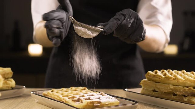 Confectioner in black gloves powdered Belgian waffles with sugar powder using sieve