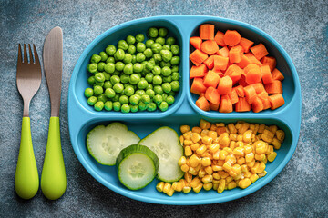 A colorful plate with green peas, diced carrots, corn, and cucumber slices, accompanied by green utensils on a textured surface.