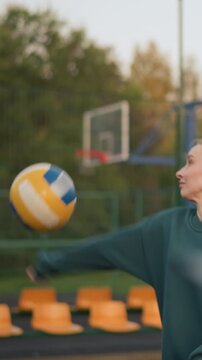 Close-up blurred view of someone observing lady in green hoodie serving volleyball on outdoor court, with background featuring people near football field and orange stadium seats
