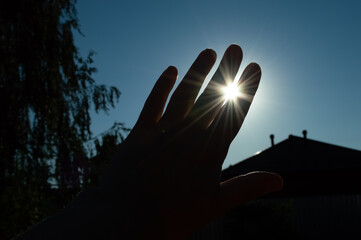 A woman's hand covers the sun at sunset, the silhouette of a house and a tree are visible