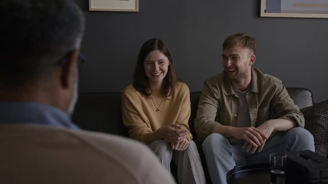 Over shoulder shot of happy young couple laughing while sitting couch together during light moment in therapy session with unrecognizable therapist listening, copy space