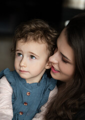 
Young mother with long brown hair holding her child with curly brown hair in a close, candid portrait indoors with a dark background